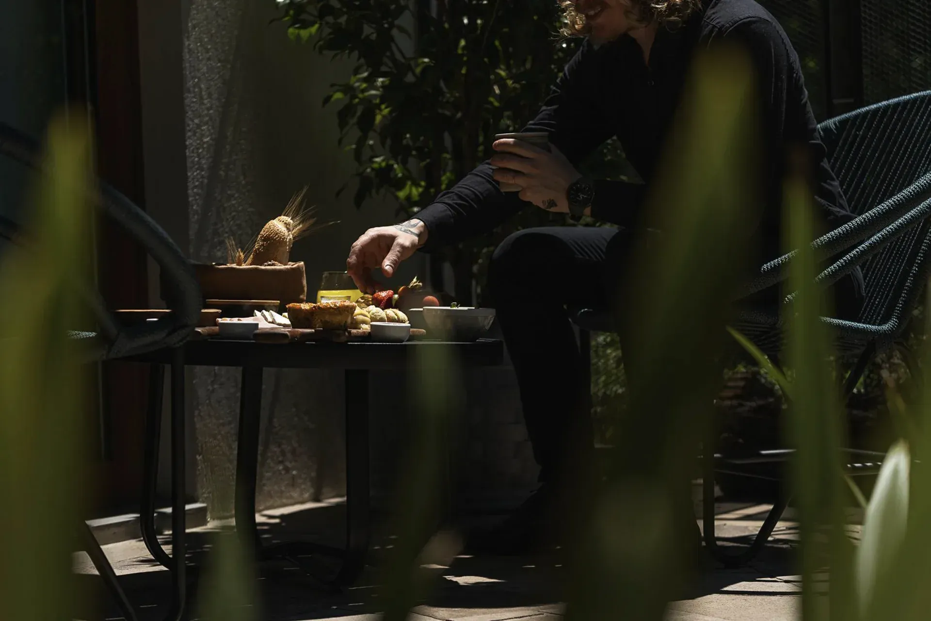 Pessoa sentada à mesa ao ar livre com comida, parcialmente escondida por plantas sob a luz solar filtrada.