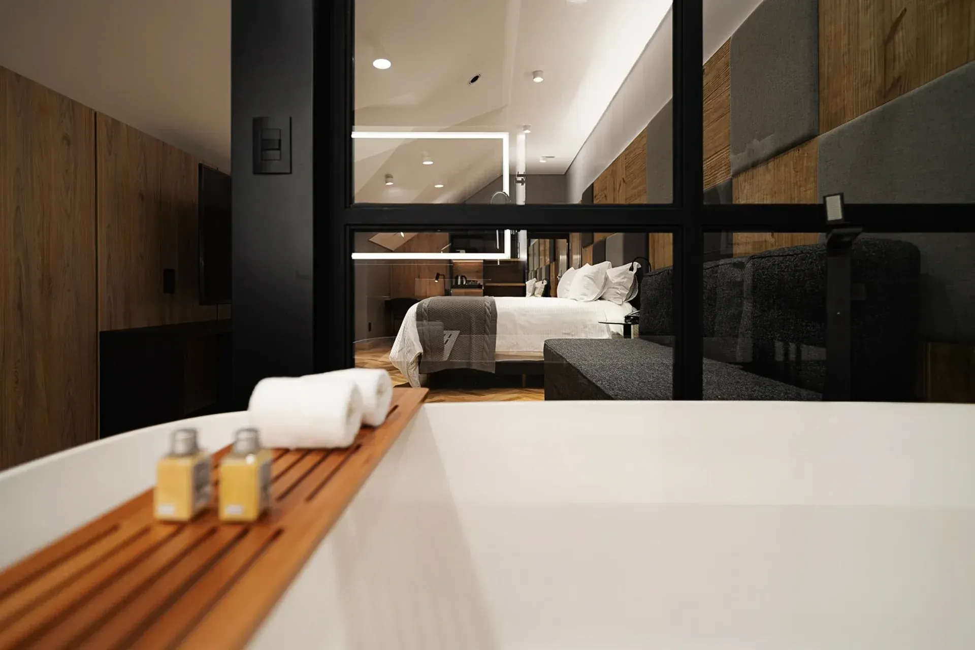 Quarto moderno com cama grande, detalhes em madeira e divisória de vidro com vista para o banheiro e banheira.