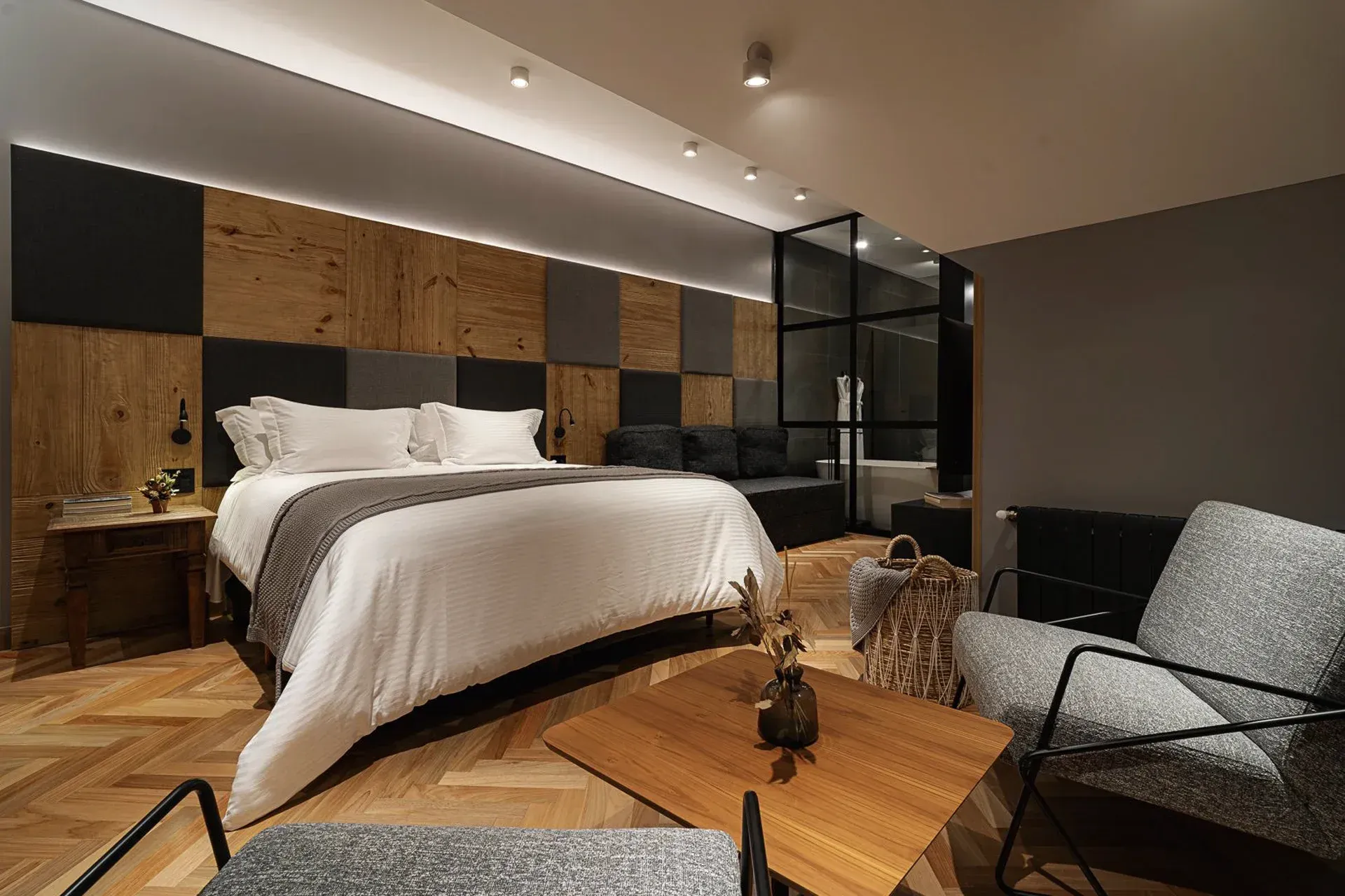 Quarto moderno com cama grande, parede com painéis de madeira, detalhes em preto e uma área de estar aconchegante.