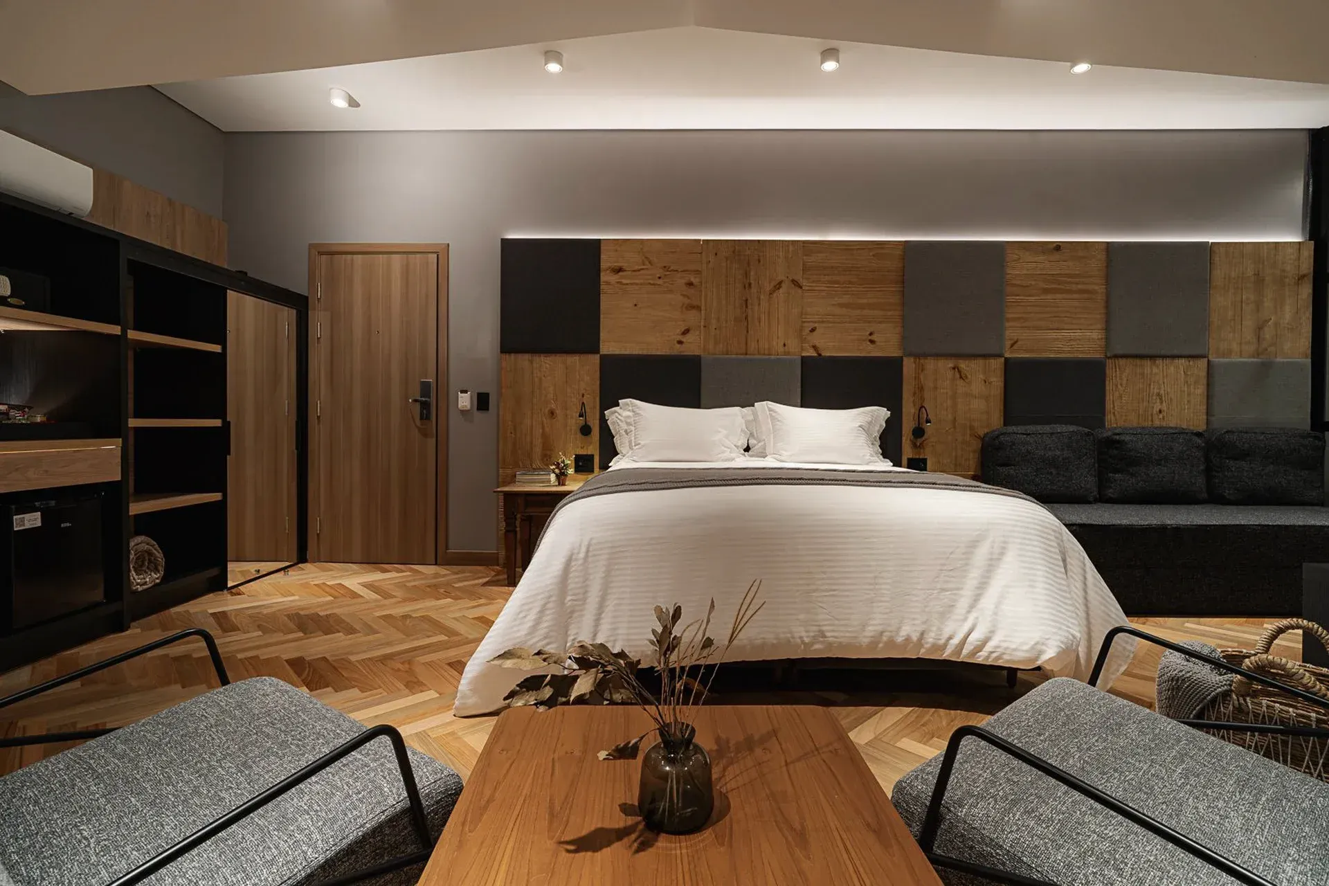 Quarto moderno com cama grande, cabeceira com painéis de madeira, prateleiras pretas e uma área de estar aconchegante.