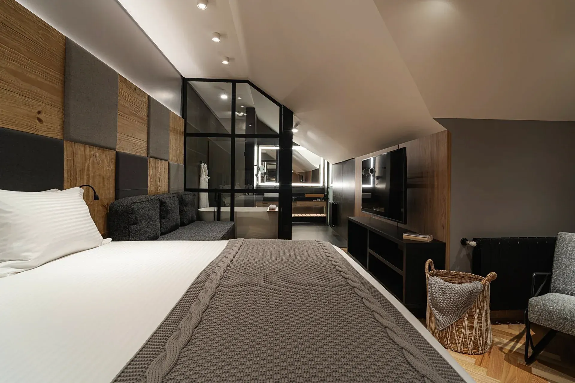 Quarto de hotel moderno com uma cama grande, divisória de vidro e decoração aconchegante em madeira e preto.