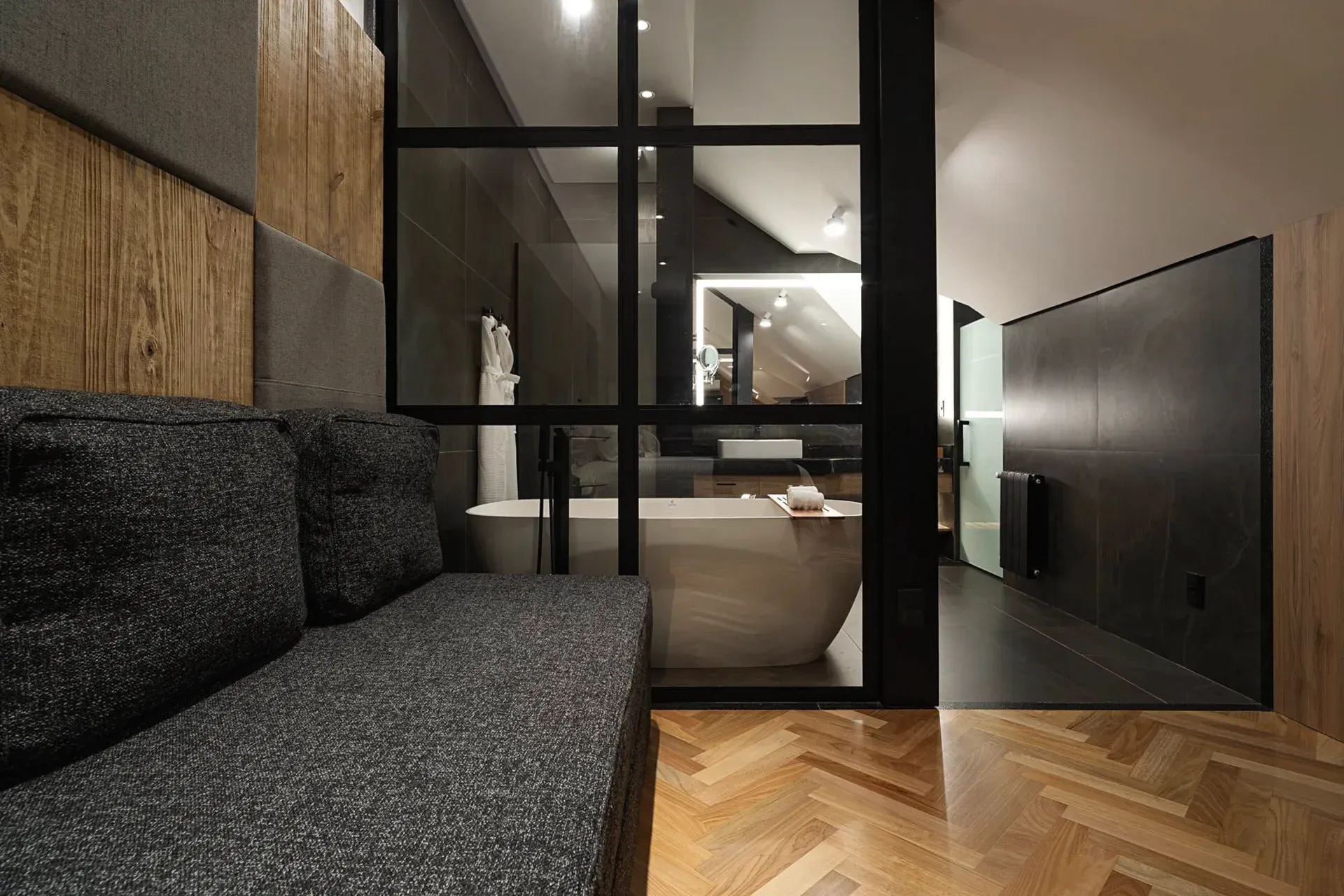 Sala de estar moderna com sofá escuro, piso de madeira em espinha de peixe e divisória de vidro.