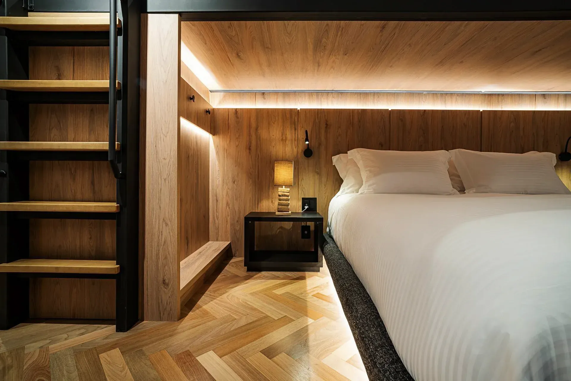 Quarto moderno com painéis de madeira, cama grande, abajur e prateleiras embutidas na escada.