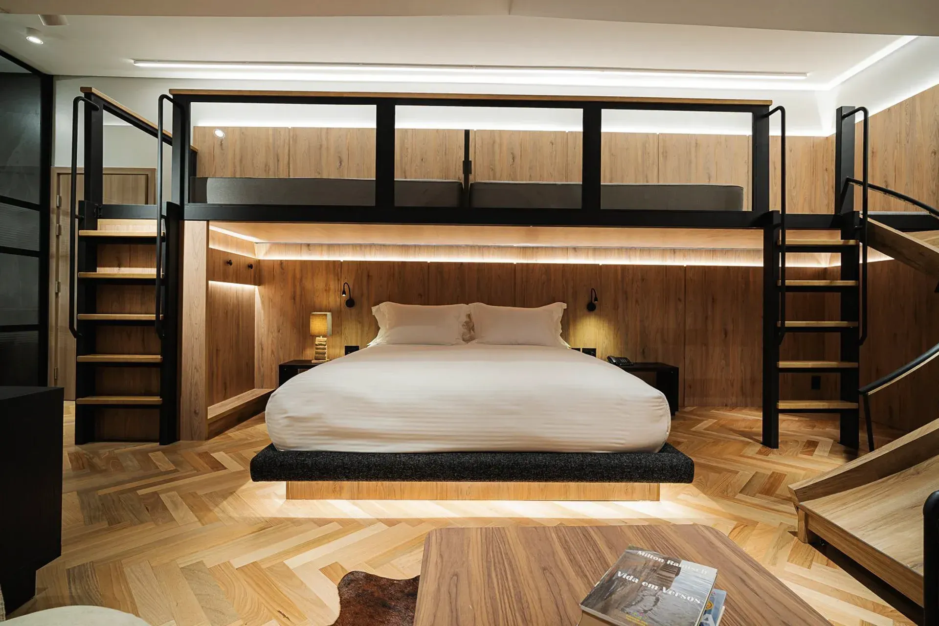 Quarto moderno em estilo loft com cama baixa, painéis de madeira em tons quentes e escadas que levam aos beliches superiores.