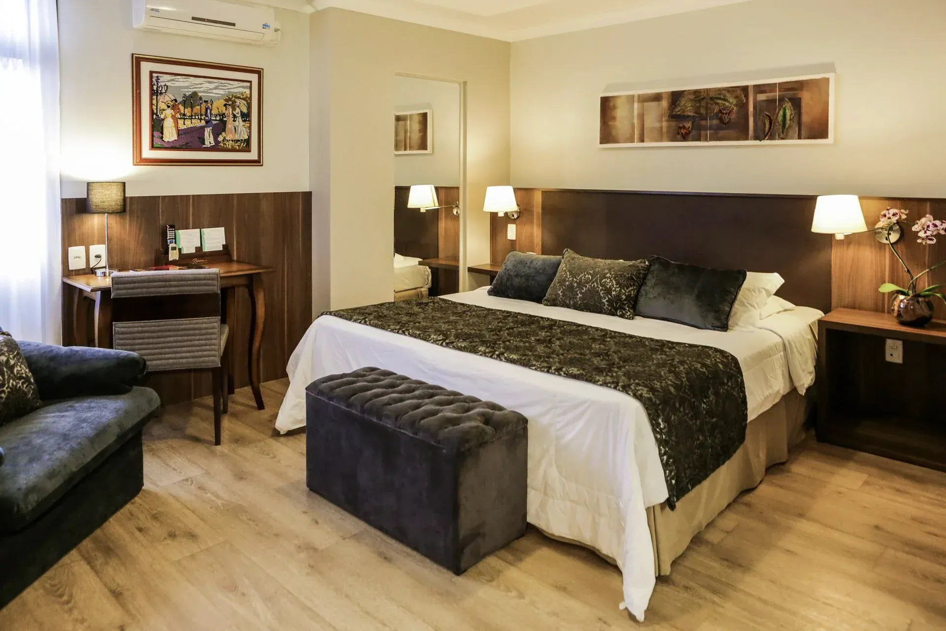Quarto de hotel bem arrumado com cama de casal, banco, luminárias, escrivaninha e sofá em tons de bege aconchegantes.