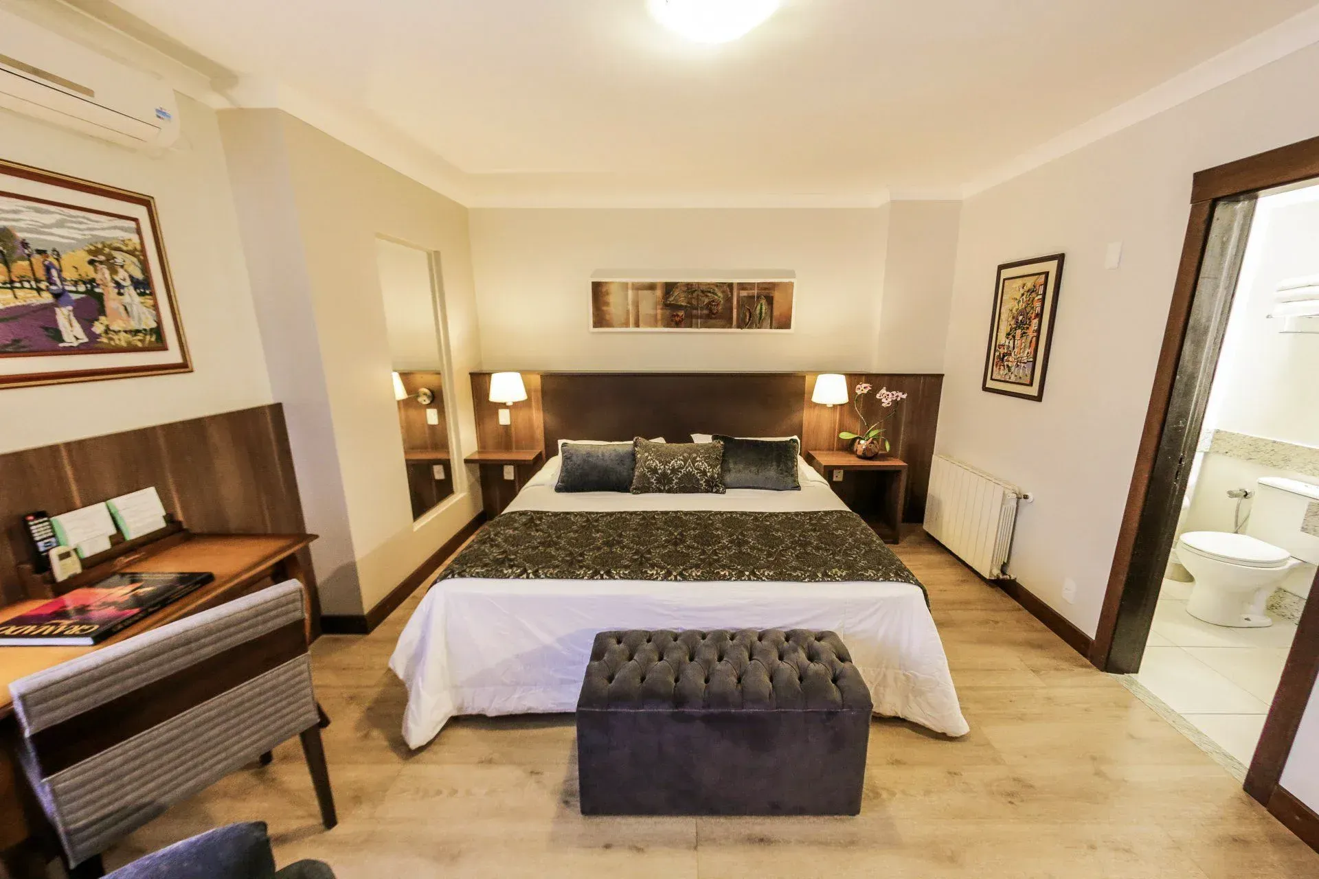 Quarto de hotel moderno com cama king-size, abajures, escrivaninha e porta de banheiro aberta.