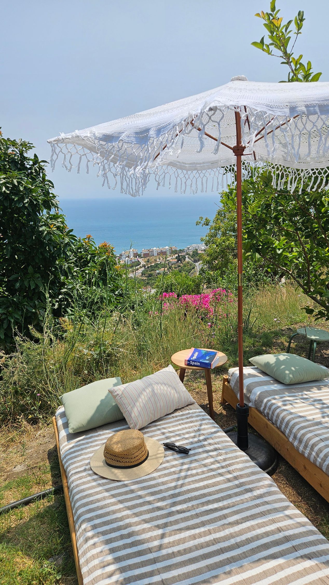 Two chaise lounges with cushions and hat under a parasol, overlooking a coastal town.
