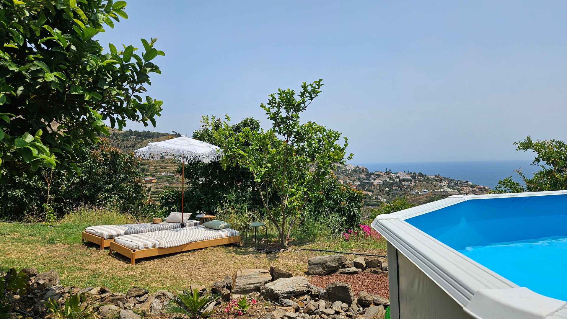 Poolside scene: blue pool, white sun umbrella, two lounge chairs, and ocean view on a sunny day.