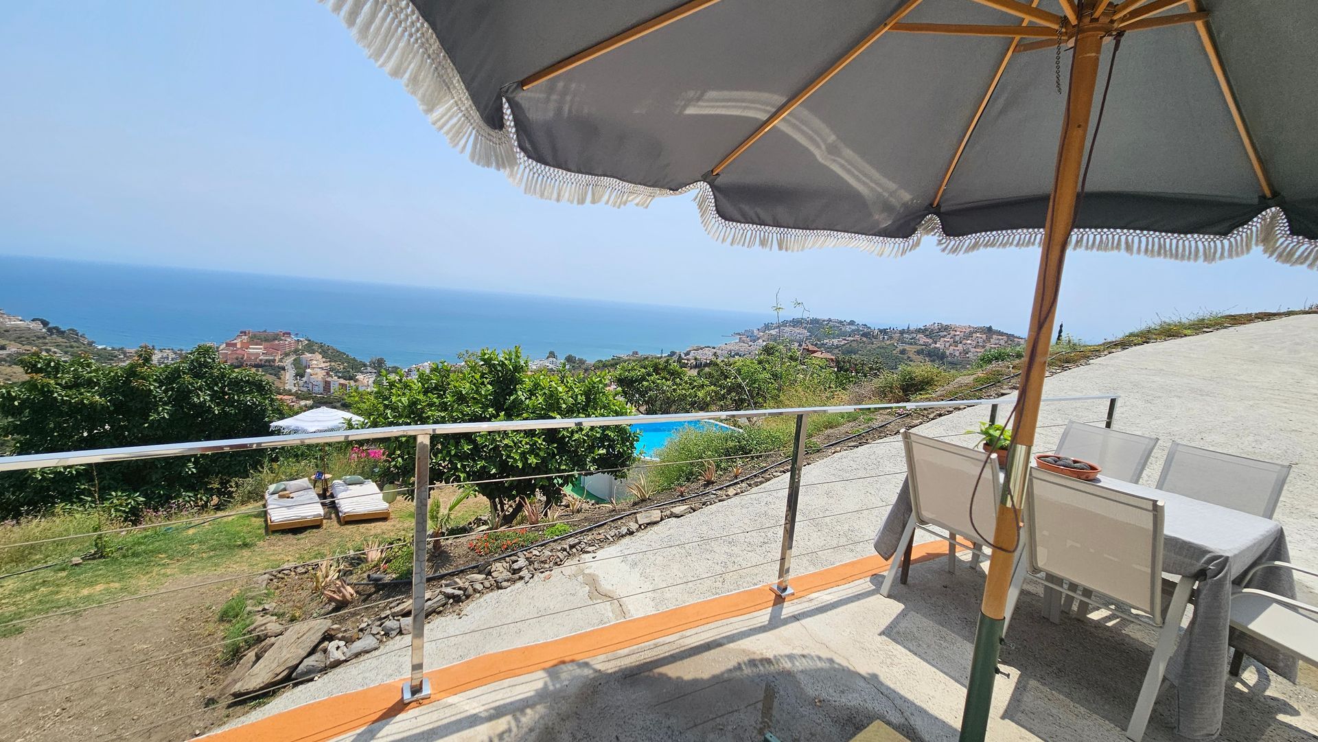 Patio overlooking the ocean with table, chairs, and umbrella. A pool and trees are visible in the distance.