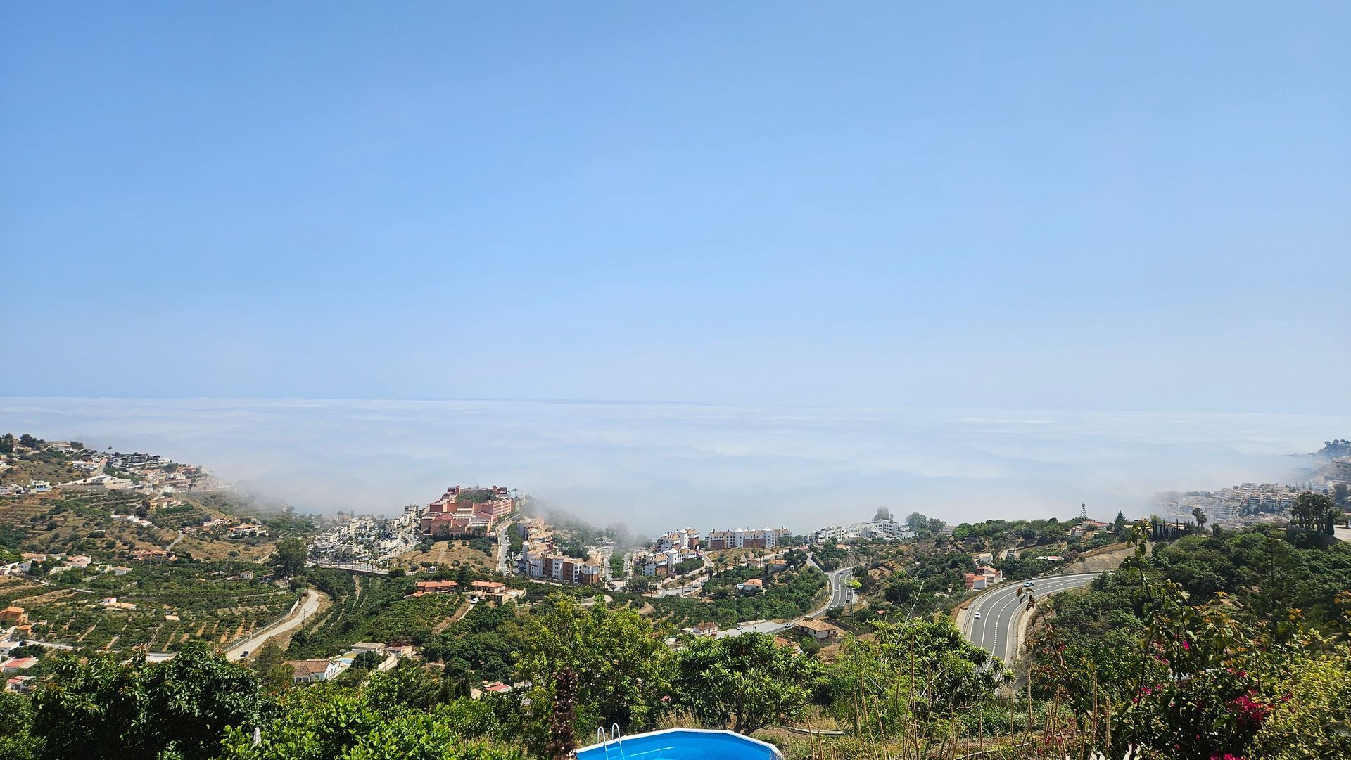 Hilly town landscape with buildings, road, and cloud-like mist; clear blue sky above.