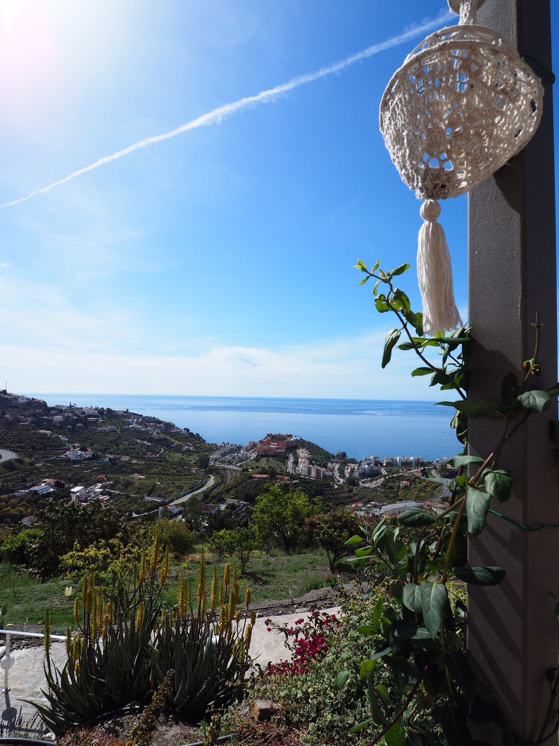 View of a coastal town and sea under a bright blue sky, with decorative hanging lamp and climbing greenery in foreground.