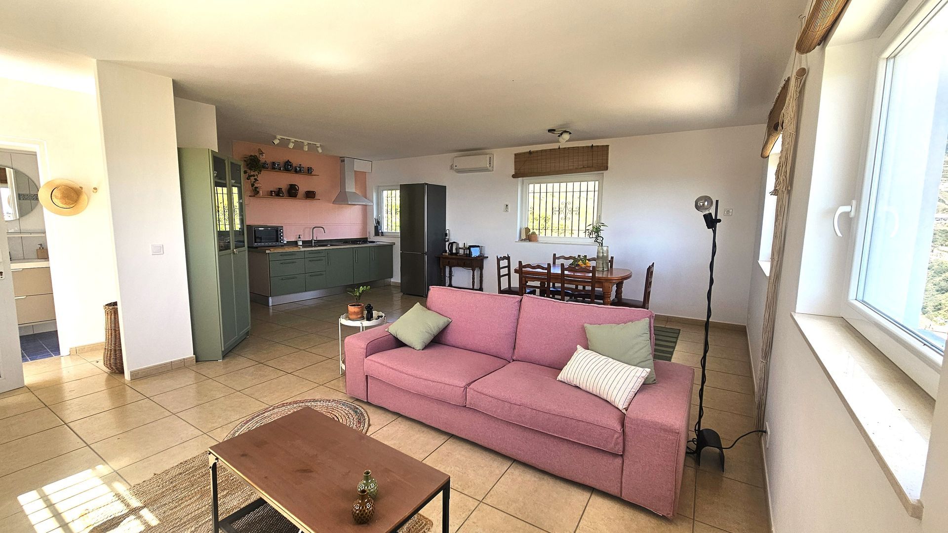Living room with pink sofa, open to a kitchen. Beige tiled floor. Bright sunlight through window.