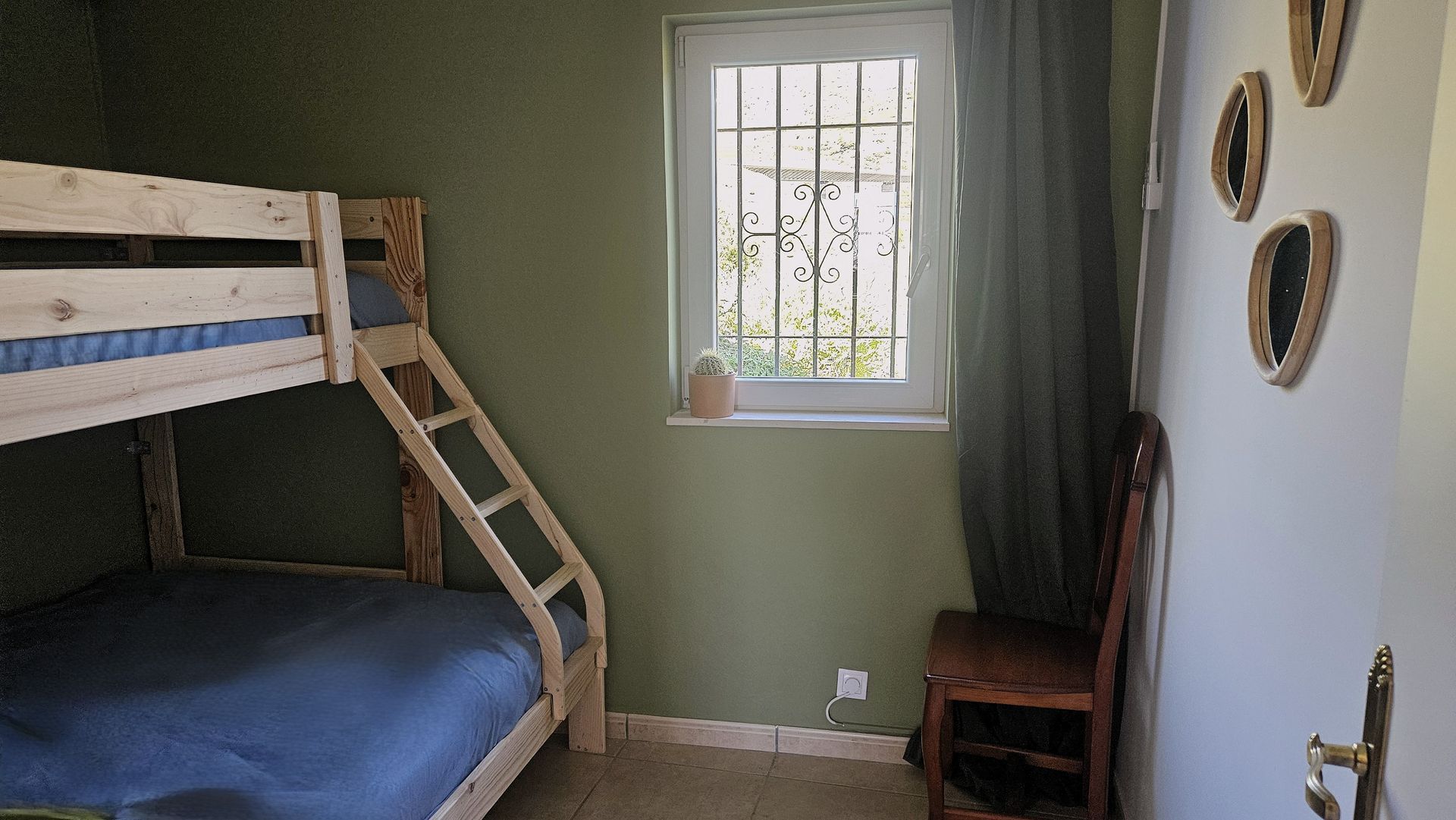 Bunk bed in a room with green walls and a window with a curtain. A chair sits by the door with decorative mirrors.