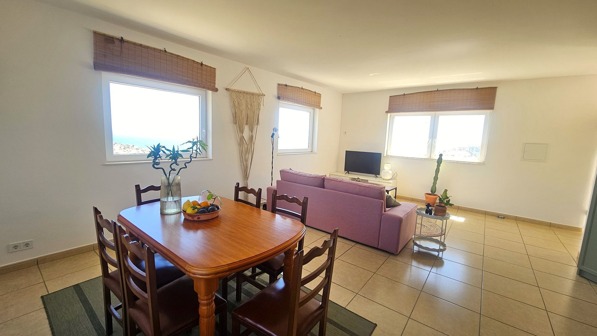 Living room with dining table, sofa, TV, and three windows overlooking the sea.