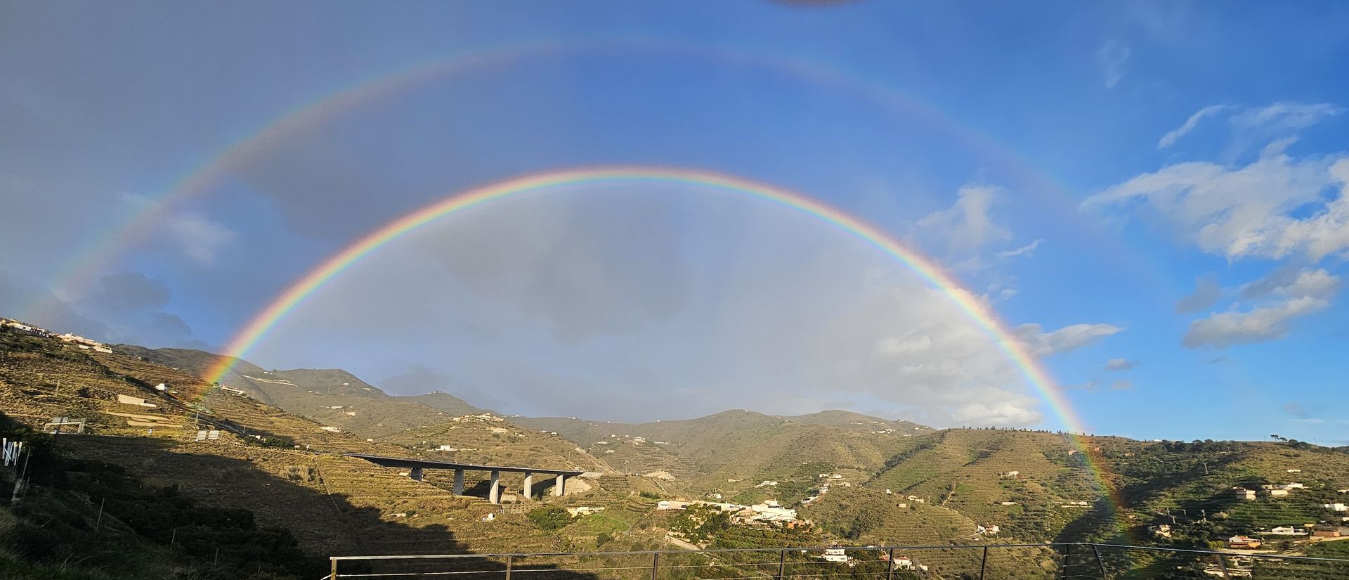 A double rainbow arcs over a mountain range under a blue sky.