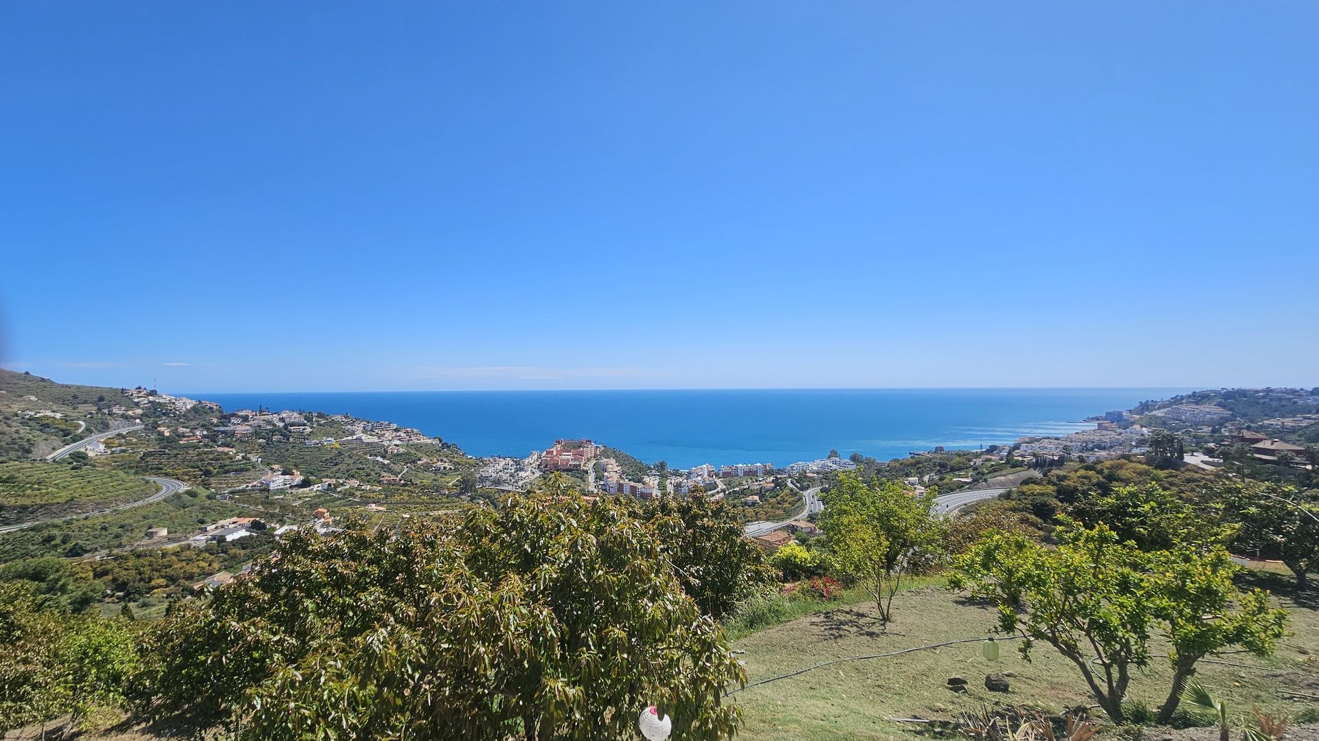 Vast ocean view from a hillside, with a clear blue sky, green vegetation, and distant town.