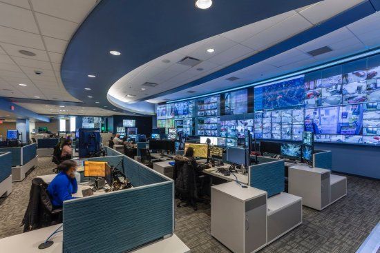 A control room with people at desks watching multiple screens, blue and gray color scheme.
