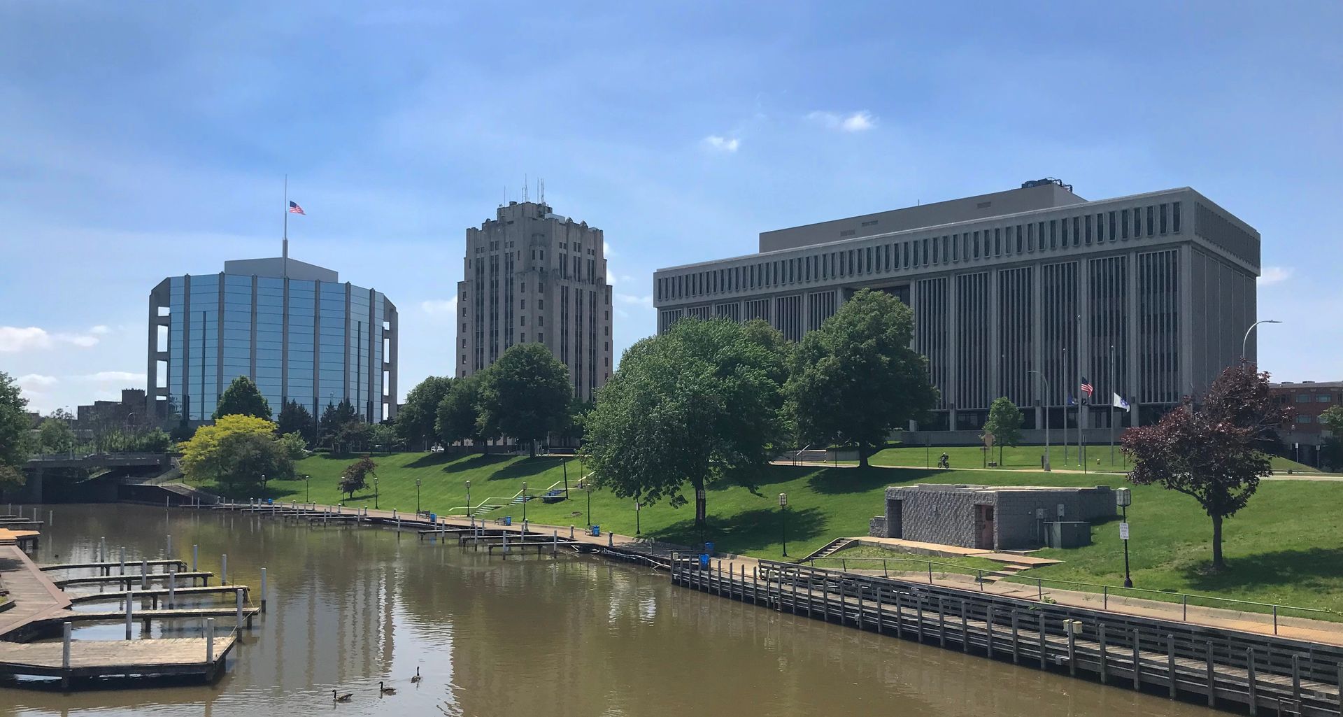 Buildings along a river, green trees, blue sky with a dock in the foreground.