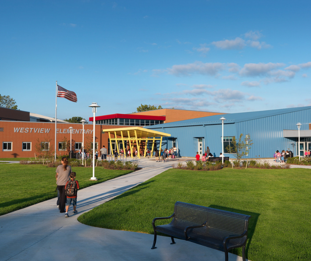 People walking to an elementary school with a blue and red exterior, grassy lawn, and American flag.