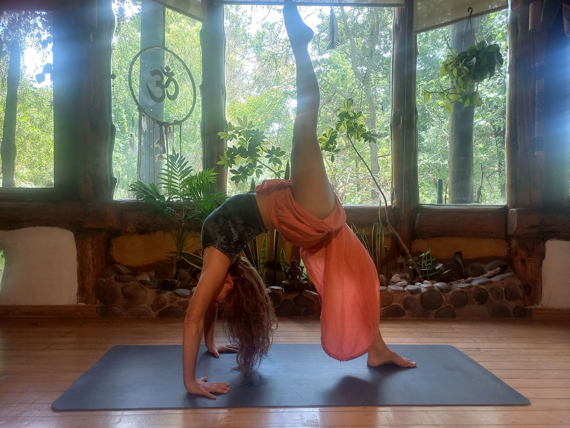 Mujer en pose de yoga aéreo, espalda arqueada, una pierna levantada, tela naranja, entorno al aire libre.
