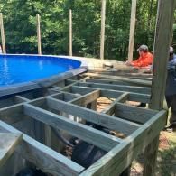 A person building a wooden deck frame next to a circular blue above-ground swimming pool in a wooded area.