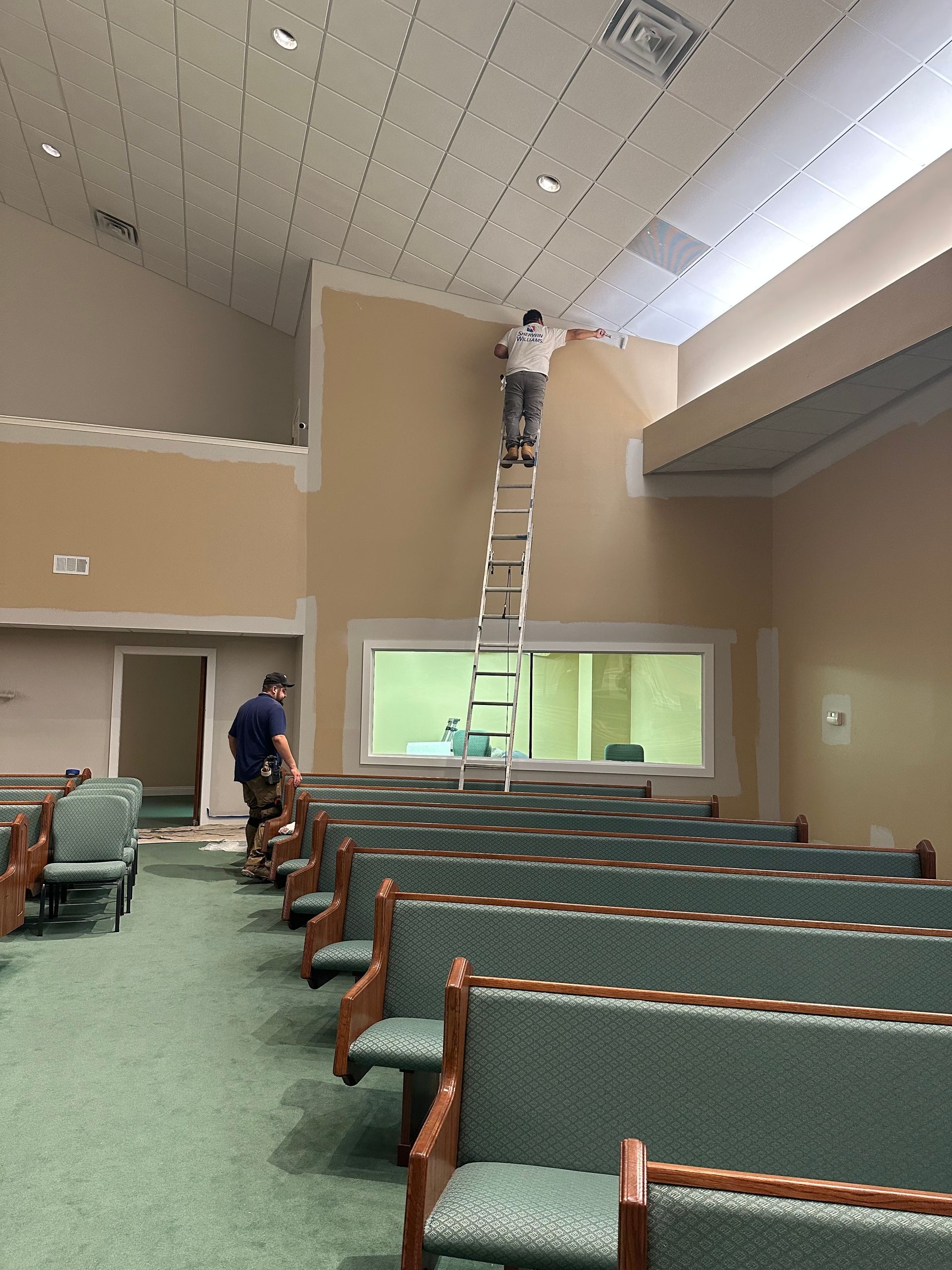 Two people work in a church with green pews; one stands on a tall ladder to paint a high wall by the ceiling.