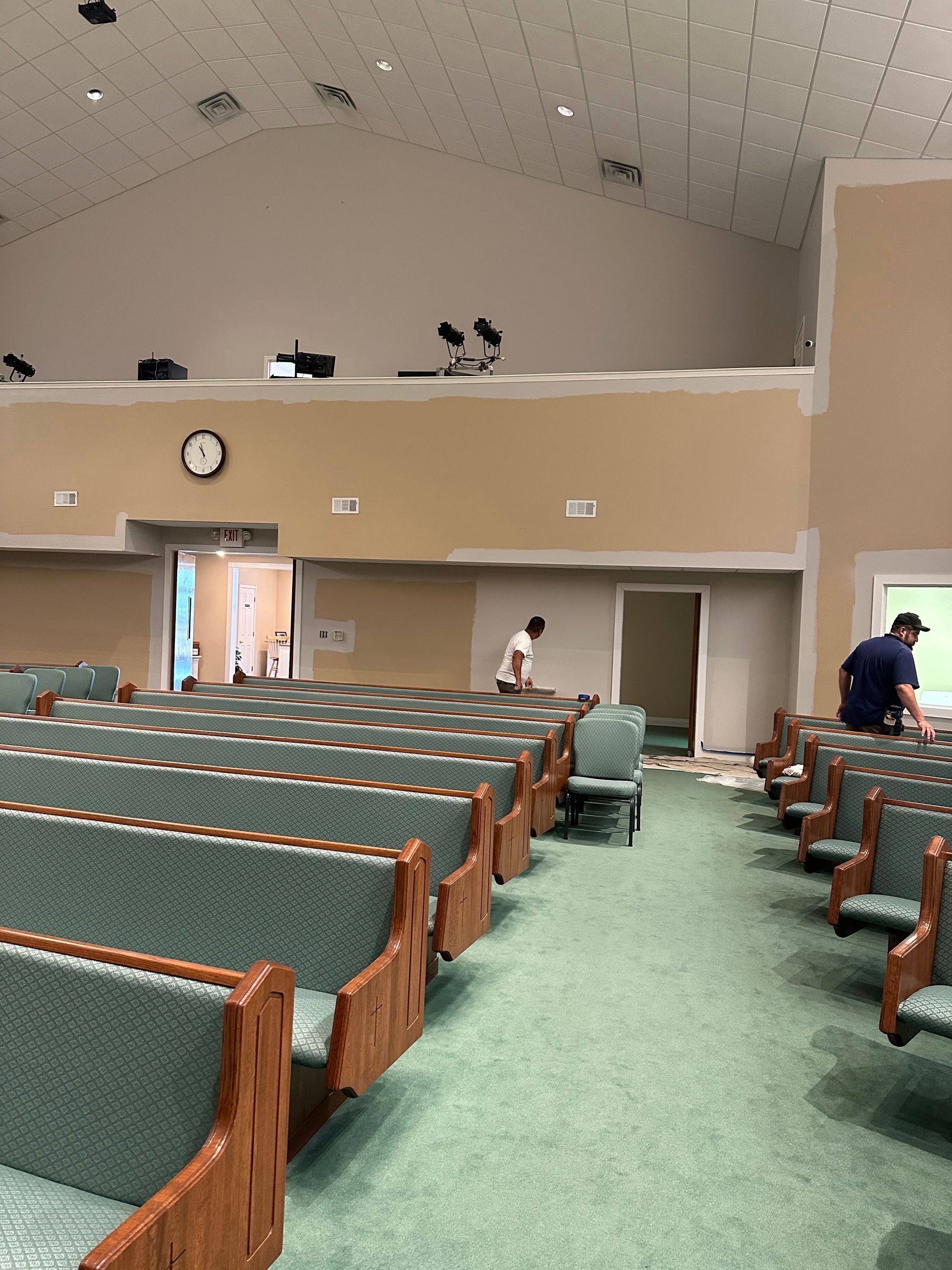 A large auditorium with rows of green-upholstered pews, beige walls under renovation, and two workers in the background.