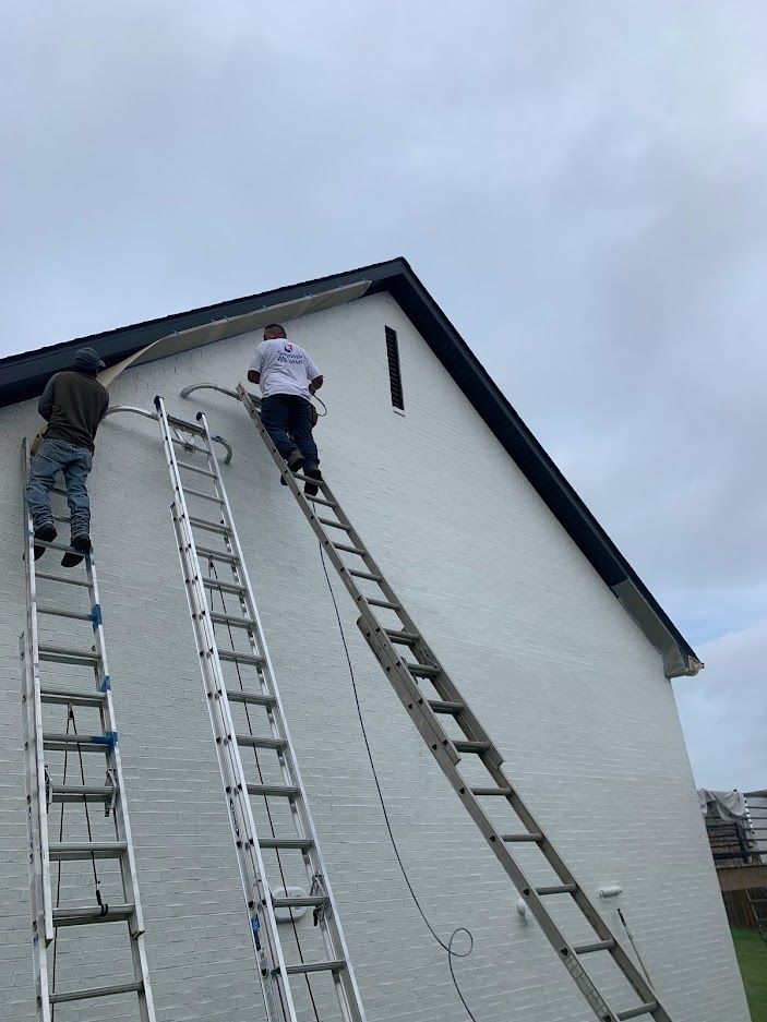 Two people on extension ladders work on the trim of a white-bricked gable end roof under a cloudy sky.