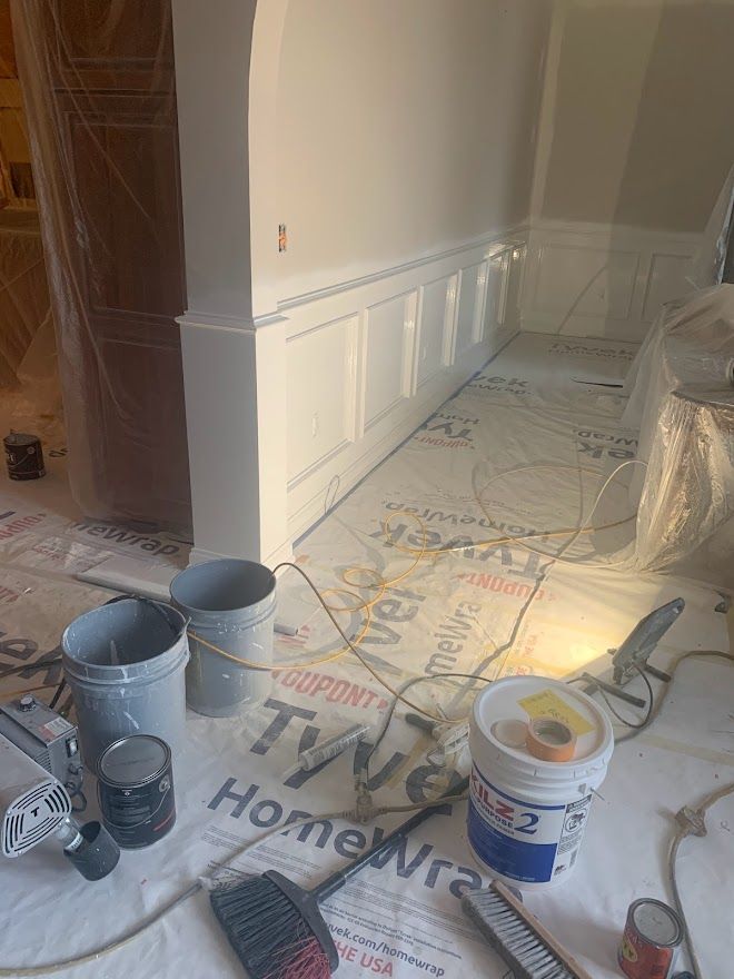 A hallway under renovation with white wainscoting, paint buckets, tools, and protective floor covering.