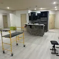 An indoor space with light flooring features a kitchen island, a yellow rolling scaffold, and a black stool.