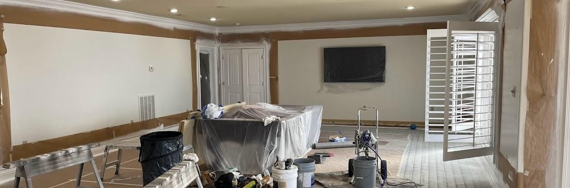 A room under renovation with taped walls, protective plastic over furniture, and tools scattered on a drop-cloth floor.