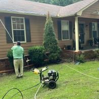 A person in a green shirt uses a pressure washer to clean the exterior of a brown house.