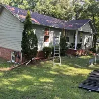 A light-colored house with a brick foundation and a metal roof, featuring a ladder placed against the side wall.