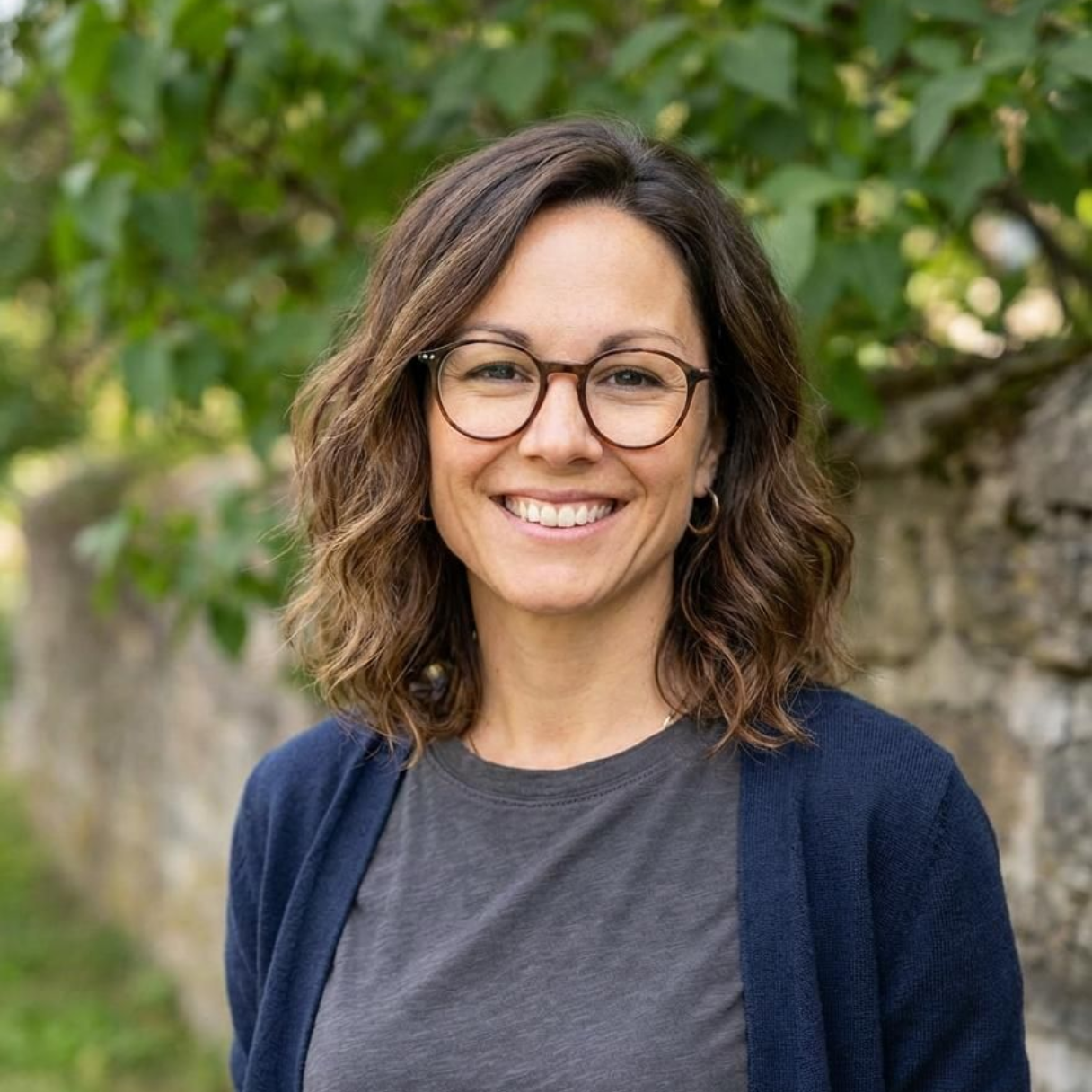 Une femme souriante portant des lunettes, vêtue d'une chemise grise et d'un cardigan bleu marine, se tient à l'extérieur, avec de la végétation verte en arrière-plan.