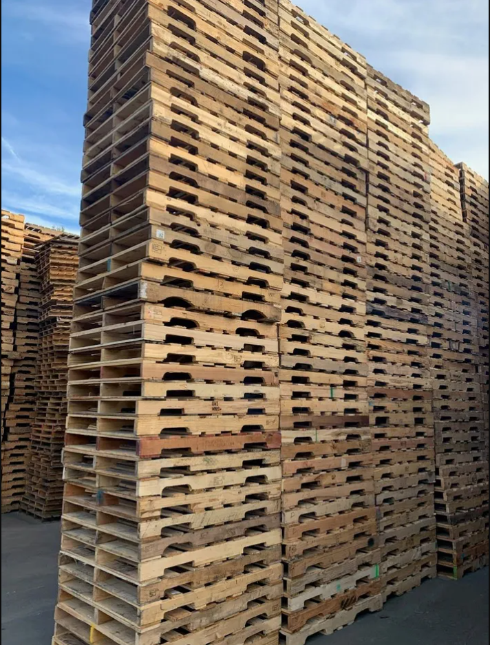 Stacks of wooden pallets stacked high outdoors on a paved lot under a partly cloudy sky