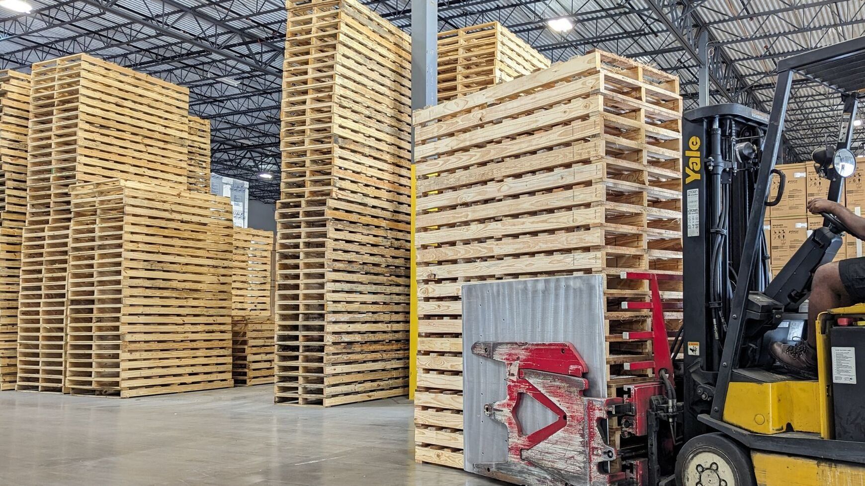 Warehouse aisle with stacked wooden pallets and a forklift carrying wrapped goods
