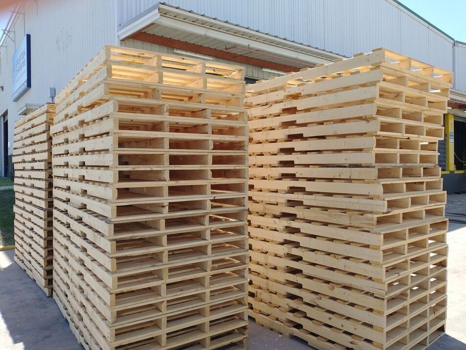 Stacks of wooden pallets beside a light-gray industrial building outdoors.