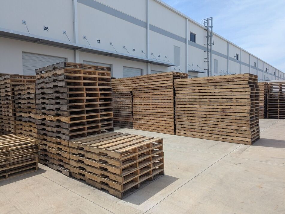 Stacks of wooden pallets in an industrial warehouse yard, beside a large gray building.