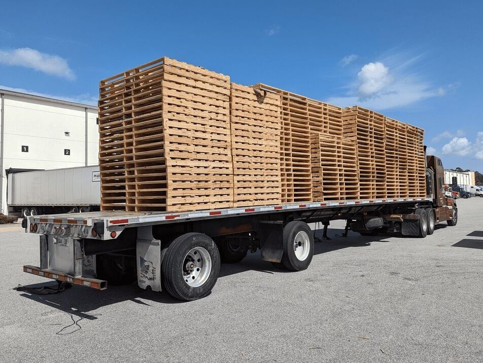 Flatbed truck loaded with stacked wooden pallets in an industrial yard under a blue sky