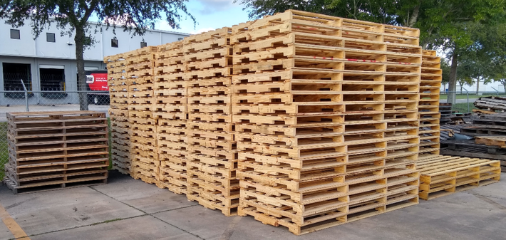Stacks of wooden pallets outdoors beside a building, with trees and pavement in the background.