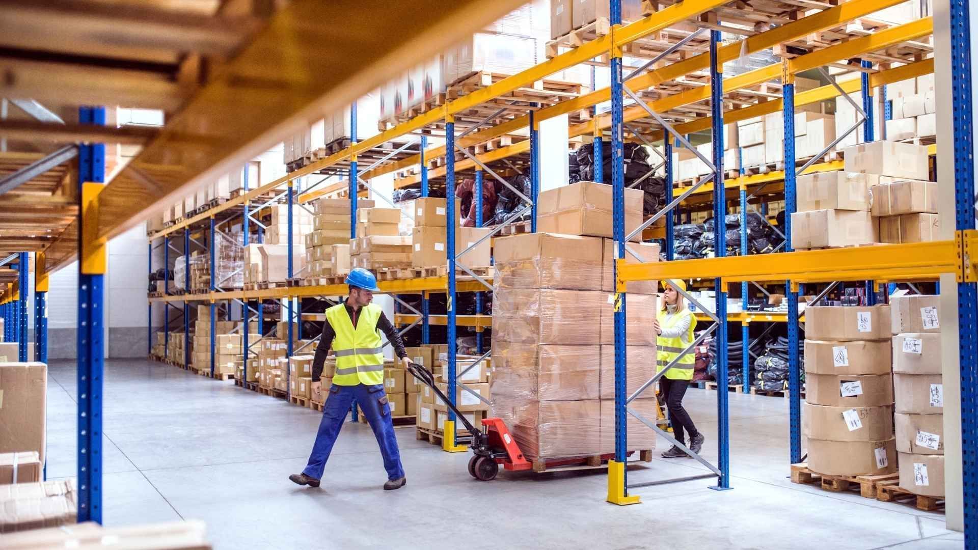Warehouse worker in a yellow vest moving boxes on a pallet jack in an industrial storage aisle