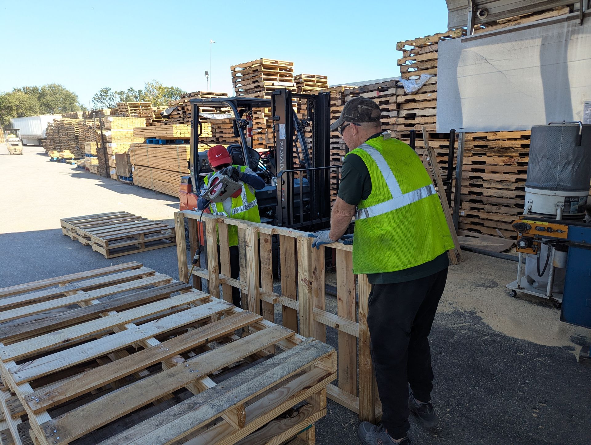 Workers in high-visibility vests moving wooden pallets in an outdoor lumber yard