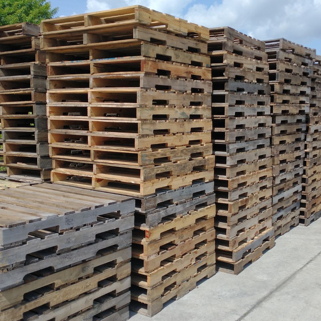 Stacks of worn wooden pallets piled beside a concrete lot outdoors.