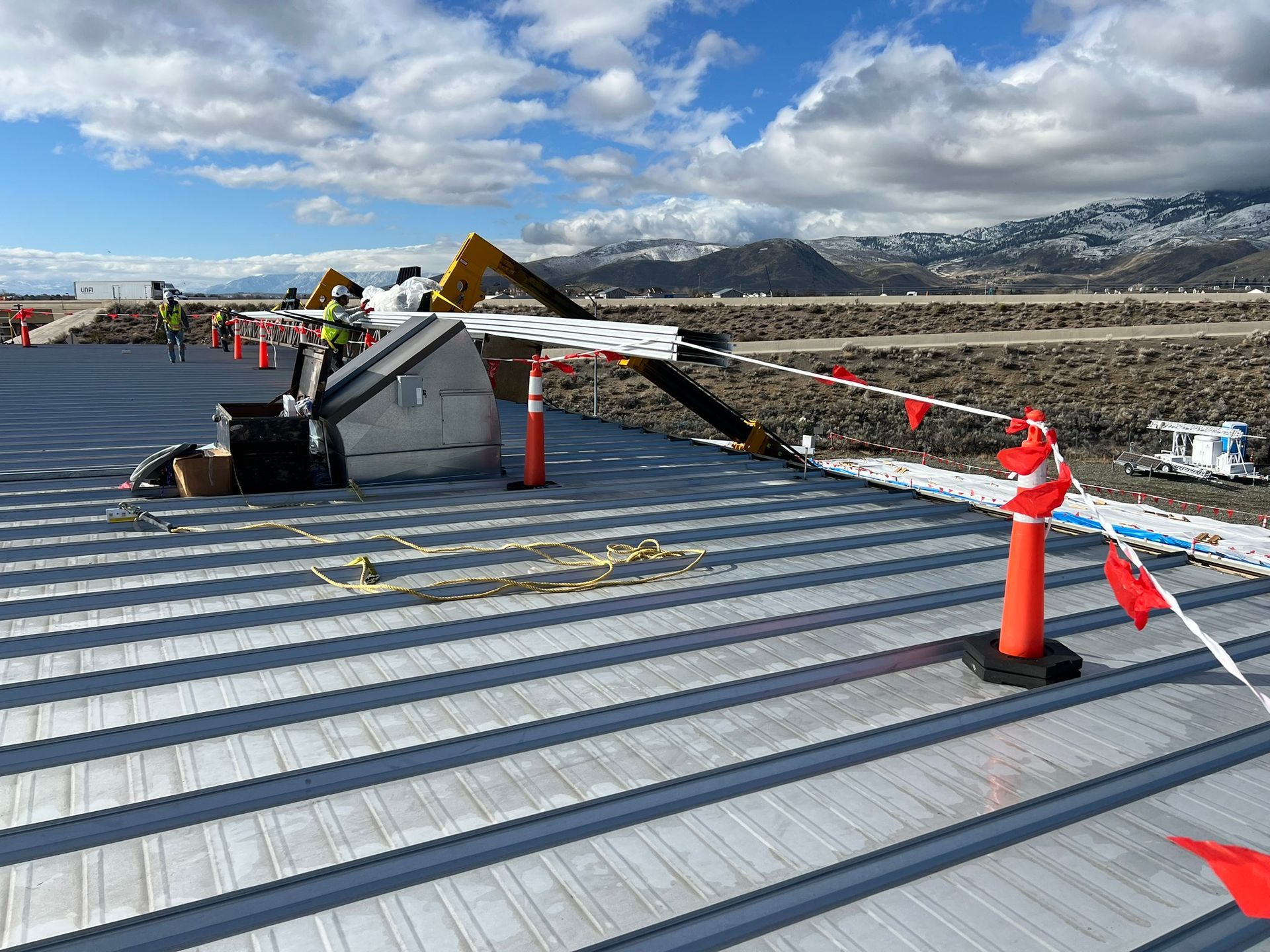 Construction site with corrugated metal roof, cones, and caution tape. Mountains in the background.