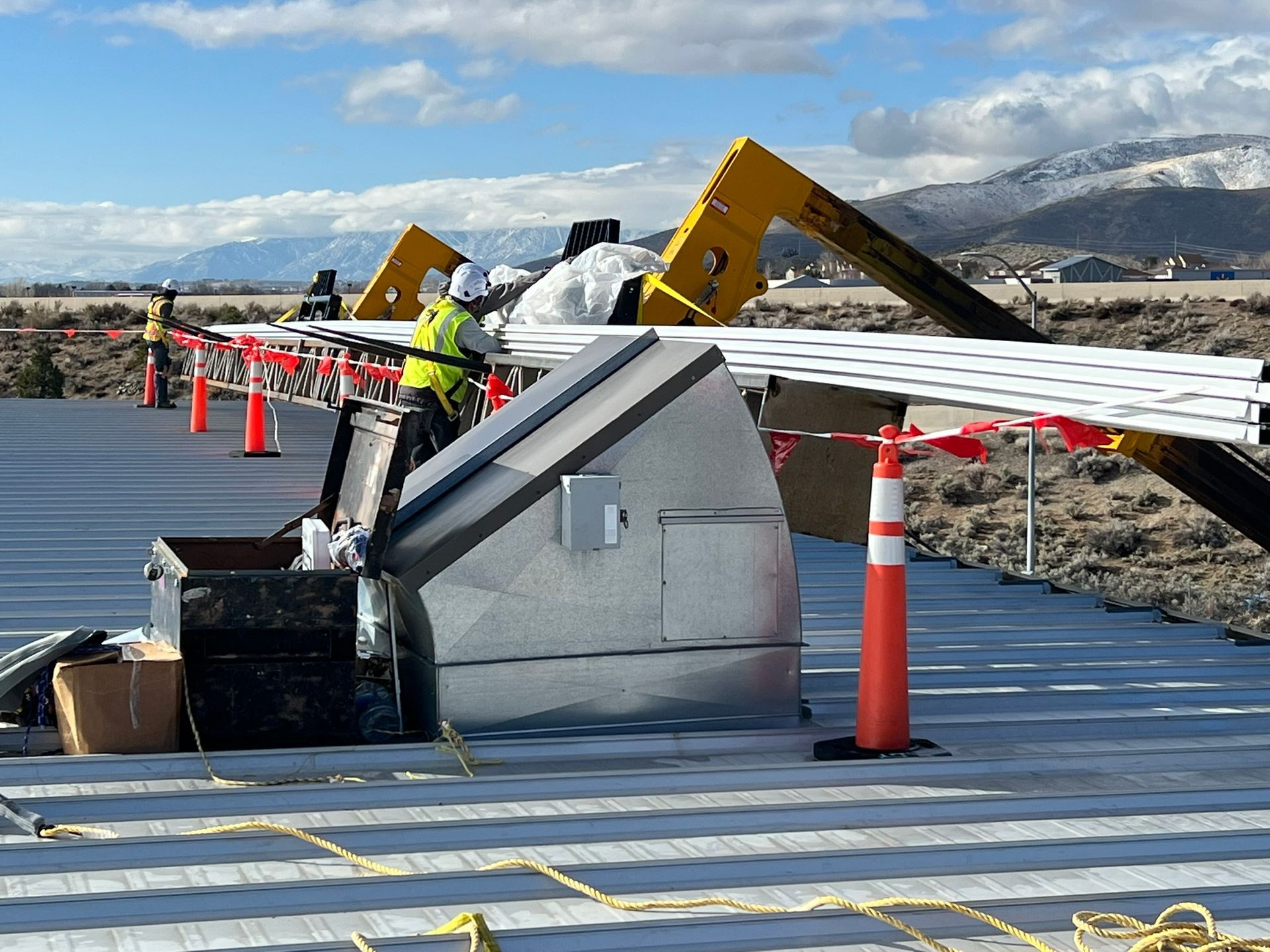 Workers in hard hats on a rooftop near a metal structure, next to a construction site in a field, under a bright sky.