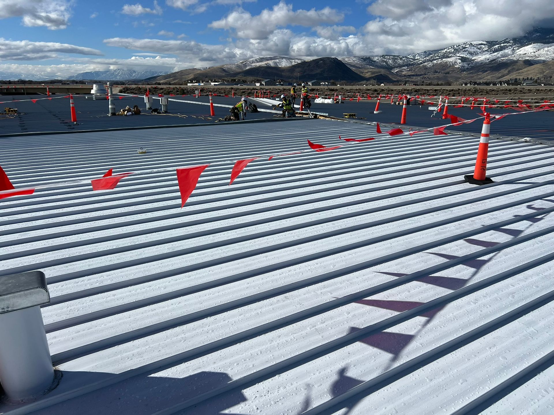 Workers on a corrugated metal roof with safety cones and flags, mountain backdrop, sunny day.