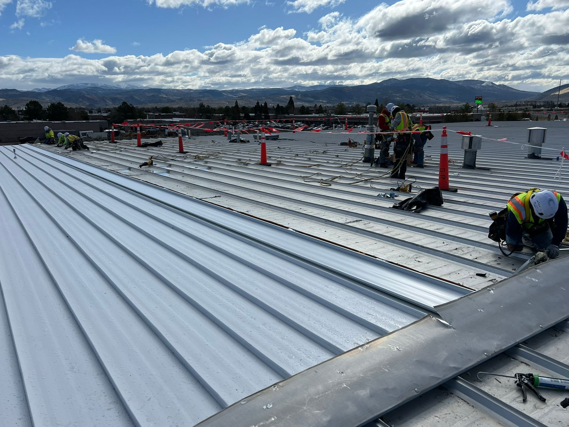 Workers installing metal roofing panels on a large rooftop, sunny day. Mountains in the background.