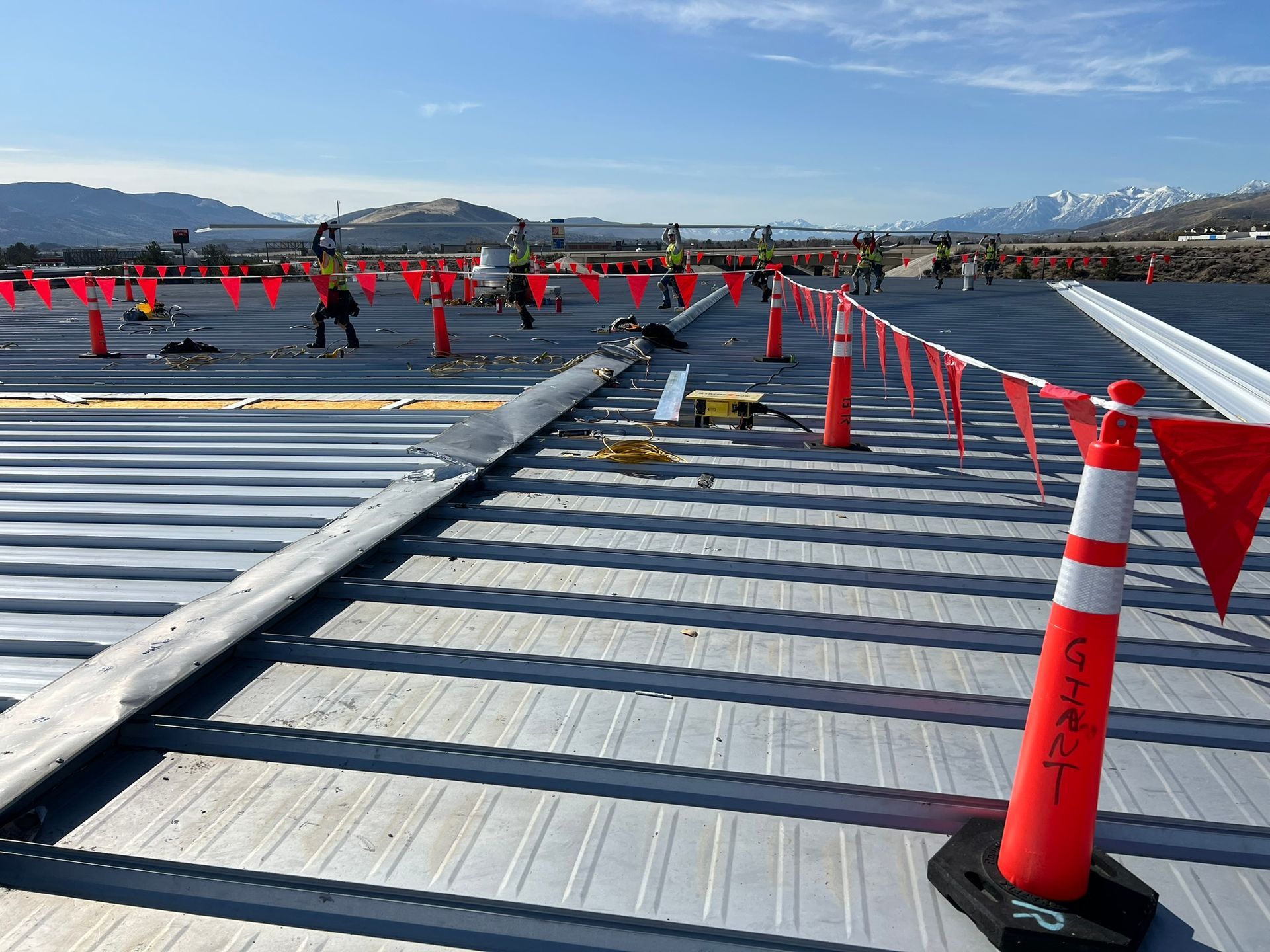 Workers installing metal roofing on a sunny day, marked by orange cones and flags. Mountains in the background.