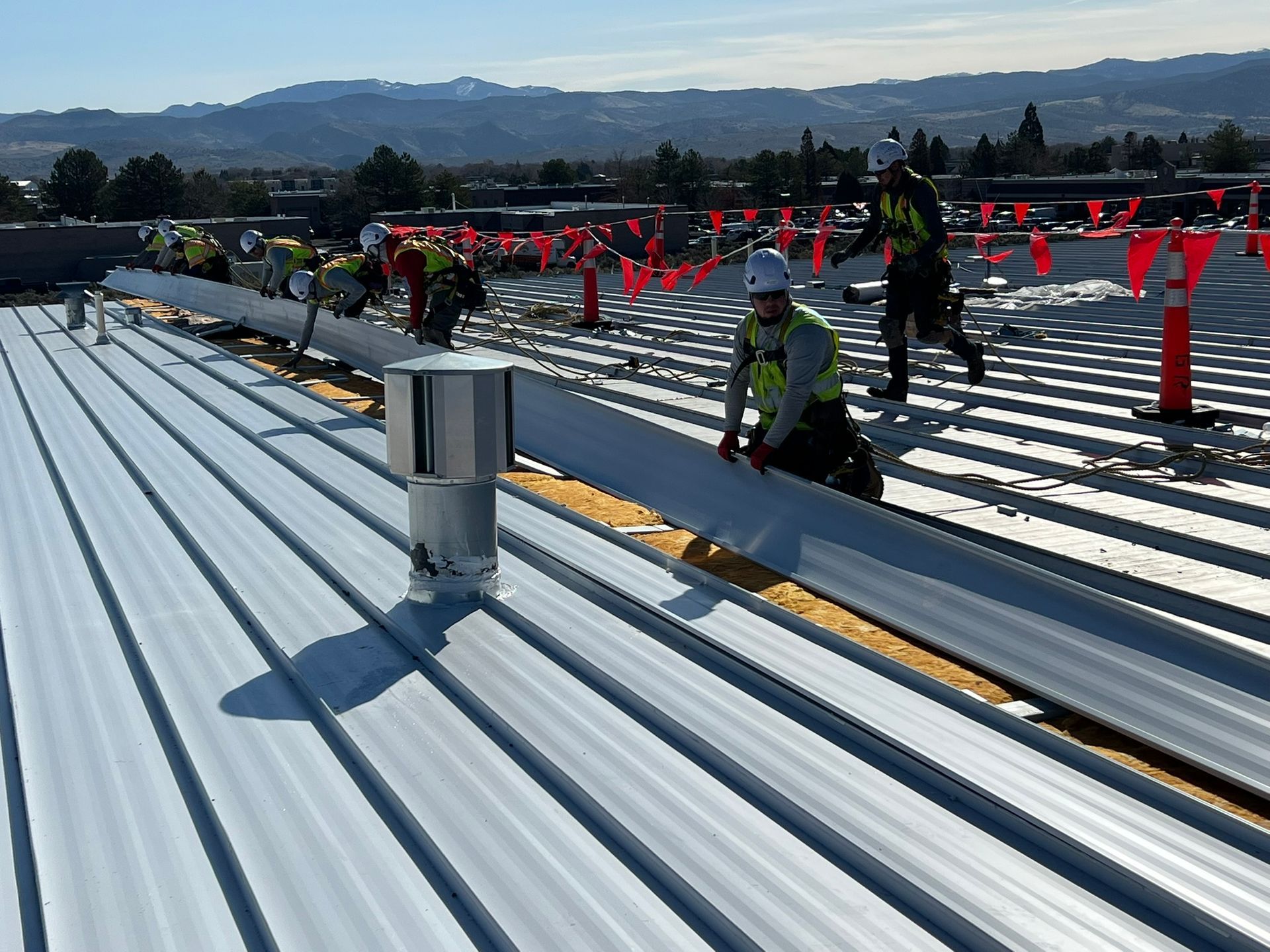 Construction workers installing metal roofing on a sunny day. Safety barriers and mountain backdrop.