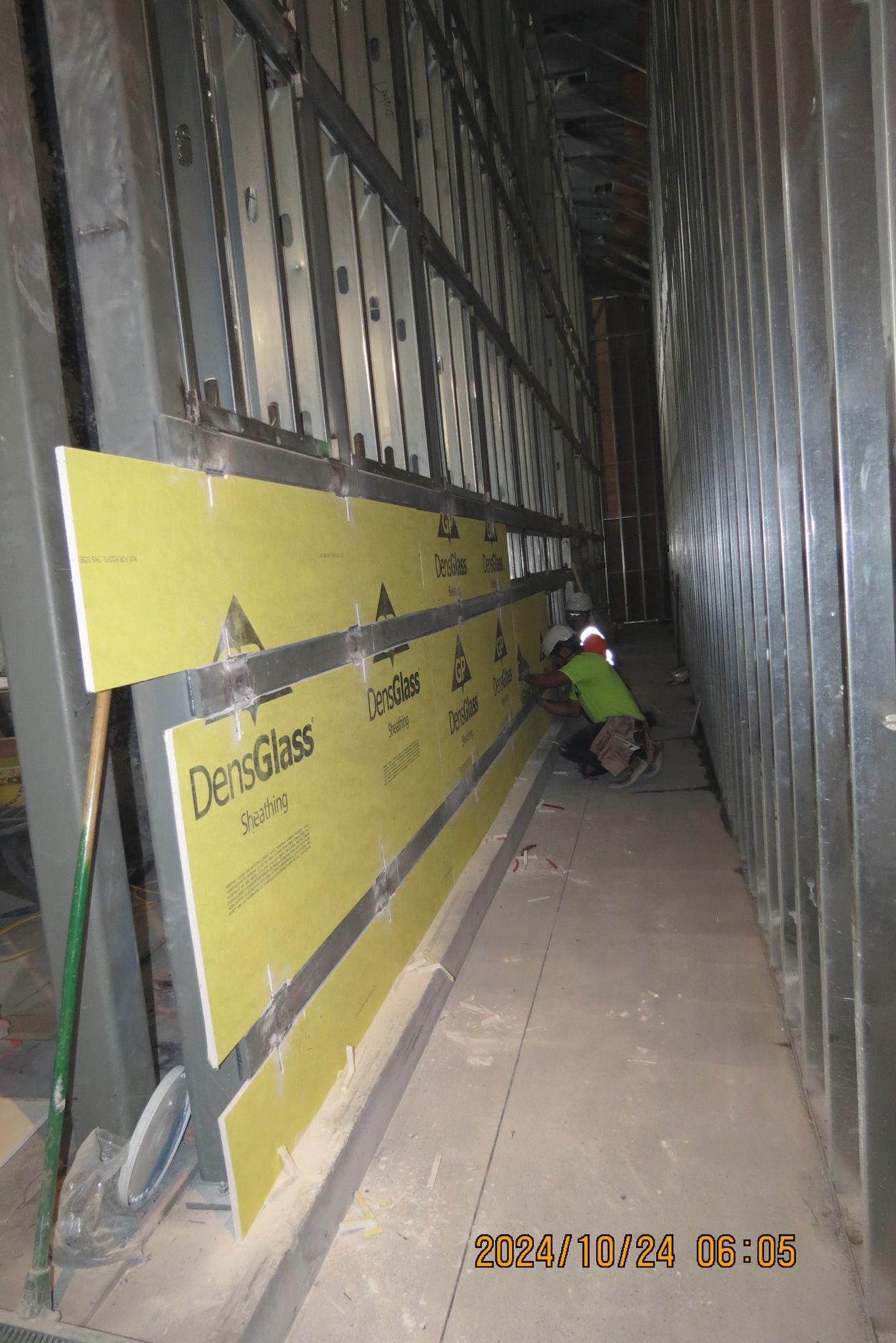 Construction worker installing yellow drywall panels between metal studs in a narrow hallway.